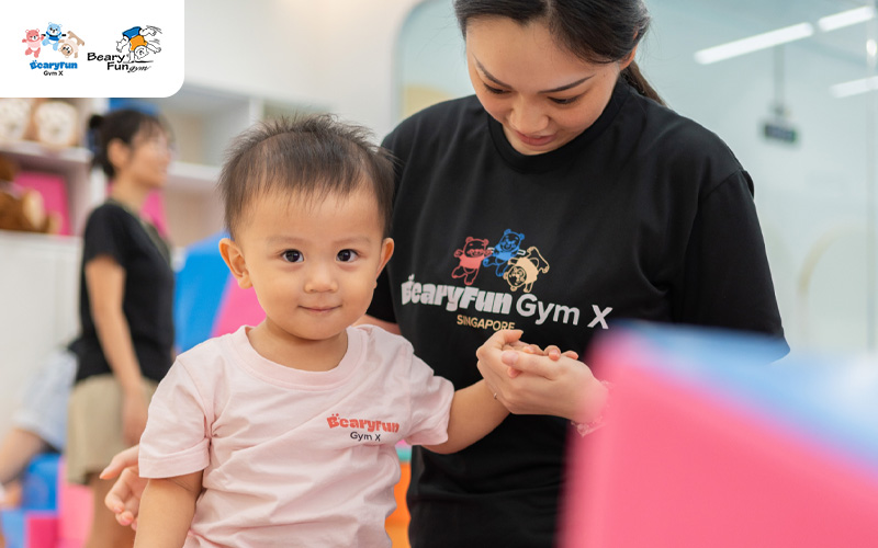 Toddler holding trainer’s hand in gym.