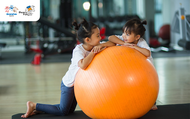 Two young girls playing with a large orange exercise ball.